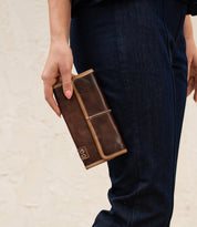 A person in dark blue jeans holds the Bed|Stü Canton, a large brown 2-in-1 leather wallet or clutch with light tan edges and a small logo on the corner.