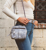 A person wearing a cream sweater and blue jeans is shown with the silver handcrafted Bed|Stü Capture purse, standing against a stone wall with a window in the background.