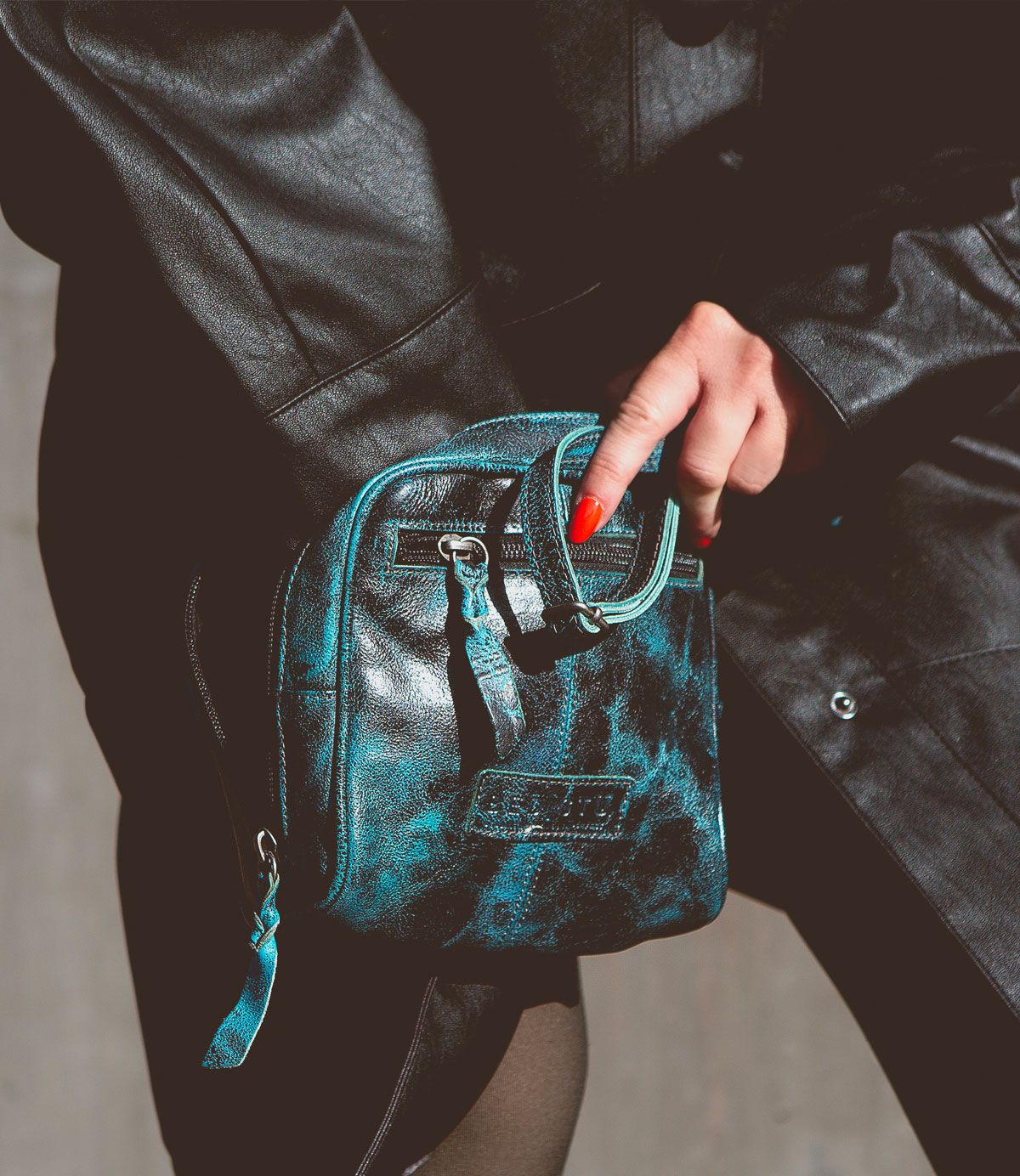 A person in a black leather coat holds the Bed|Stü Capture—a small, black and teal marbled handcrafted purse featuring a zipper and front pocket.