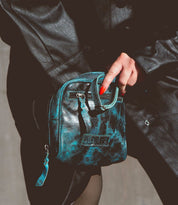 A person in a black leather coat holds the Bed|Stü Capture—a small, black and teal marbled handcrafted purse featuring a zipper and front pocket.