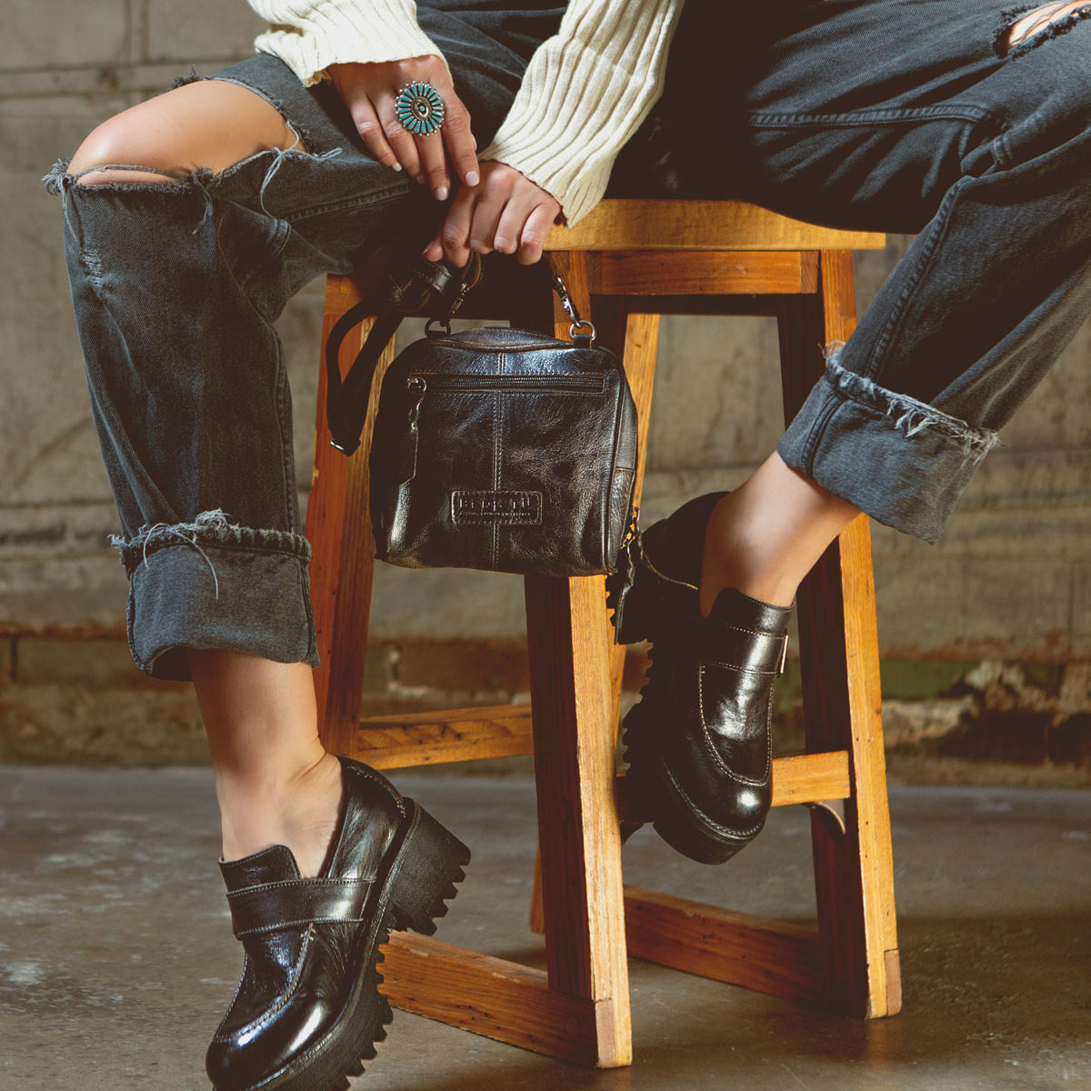 A person wearing distressed jeans, a white ribbed sweater, and black chunky loafers sits on a wooden stool, holding the Bed|Stü Capture compact handbag and displaying a turquoise ring.