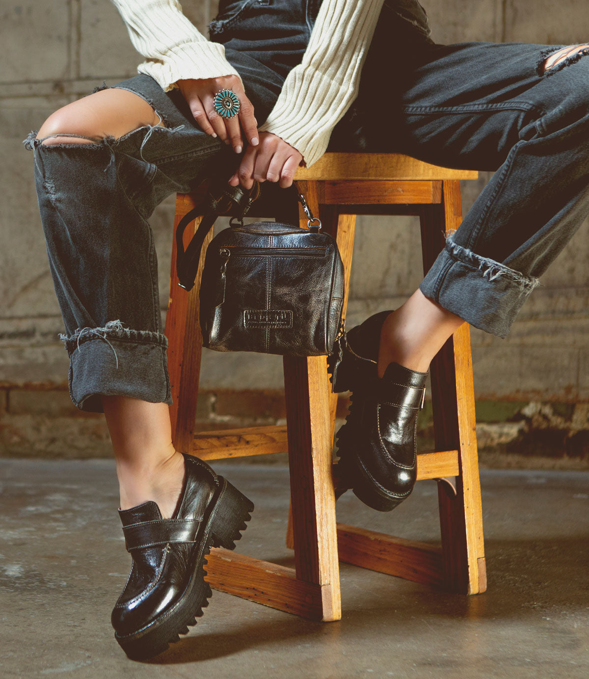 A person wearing distressed jeans, a white ribbed sweater, and black chunky loafers sits on a wooden stool, holding the Bed|Stü Capture compact handbag and displaying a turquoise ring.