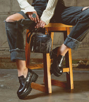 A person wearing distressed jeans, a white ribbed sweater, and black chunky loafers sits on a wooden stool, holding the Bed|Stü Capture compact handbag and displaying a turquoise ring.