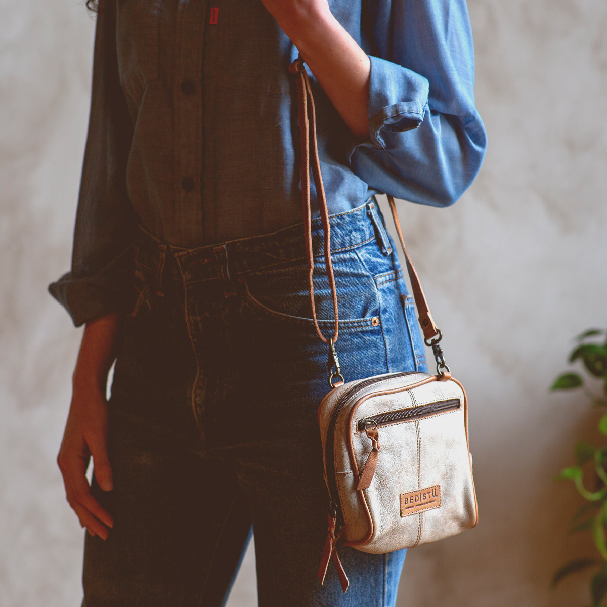 Wearing a denim shirt and jeans, the person stands indoors near a plant, holding the Bed|Stü "Capture"—a small handcrafted purse featuring a convenient zipper pocket.