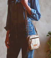 Wearing a denim shirt and jeans, the person stands indoors near a plant, holding the Bed|Stü "Capture"—a small handcrafted purse featuring a convenient zipper pocket.