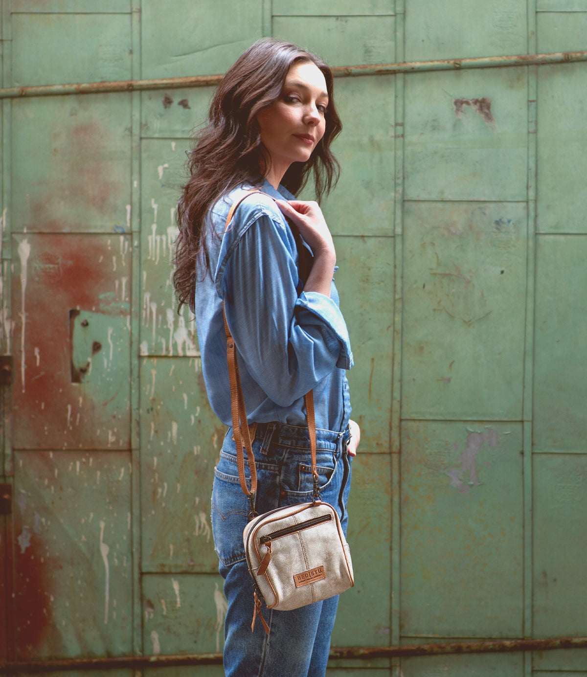 A woman in a blue denim shirt and jeans stands sideways in front of a green metal wall, carrying the small handcrafted "Capture" purse by Bed|Stü.