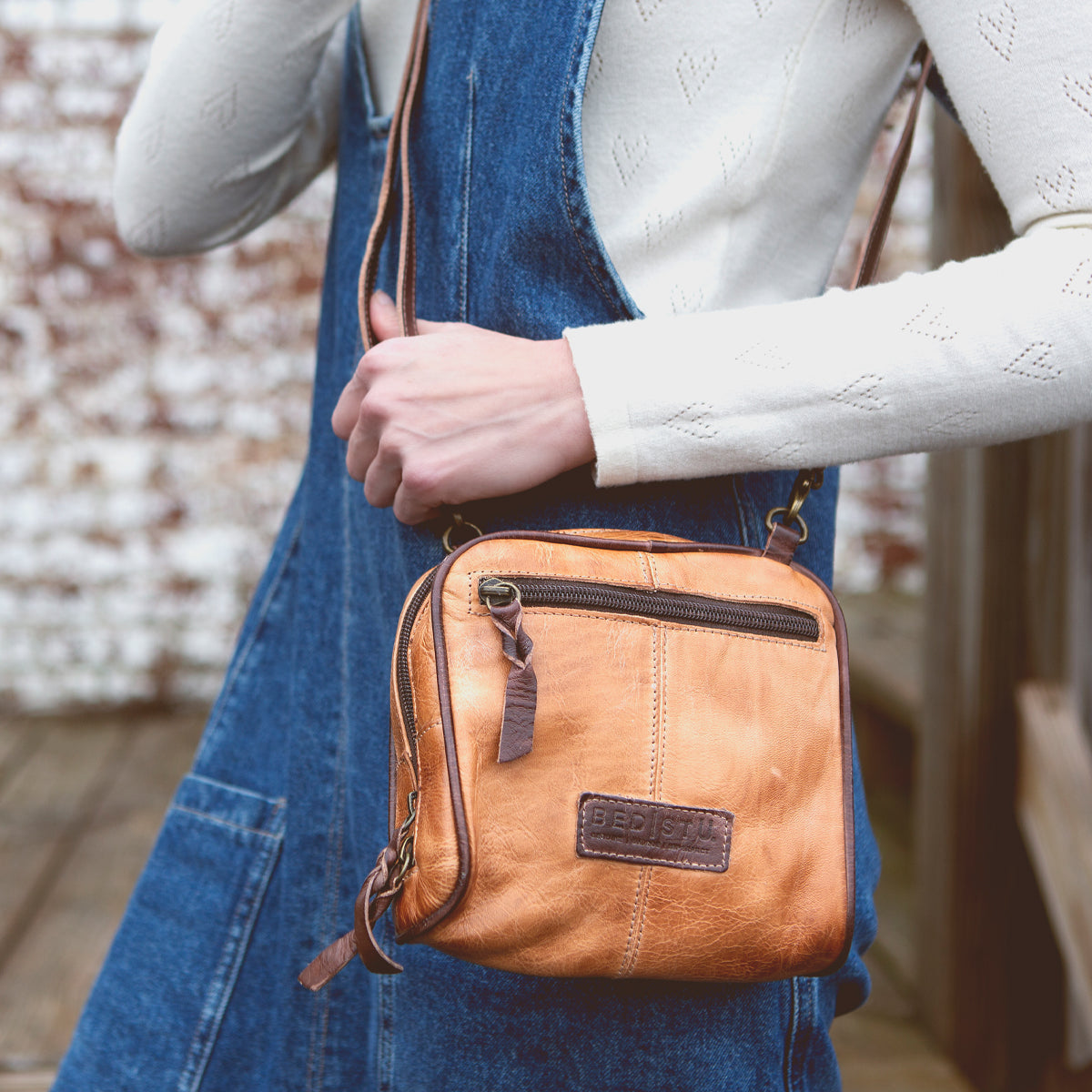A person in a white long-sleeve top and blue denim pinafore holds the Bed|Stü Capture compact handbag in tan, featuring a front zipper pocket and embossed patch.