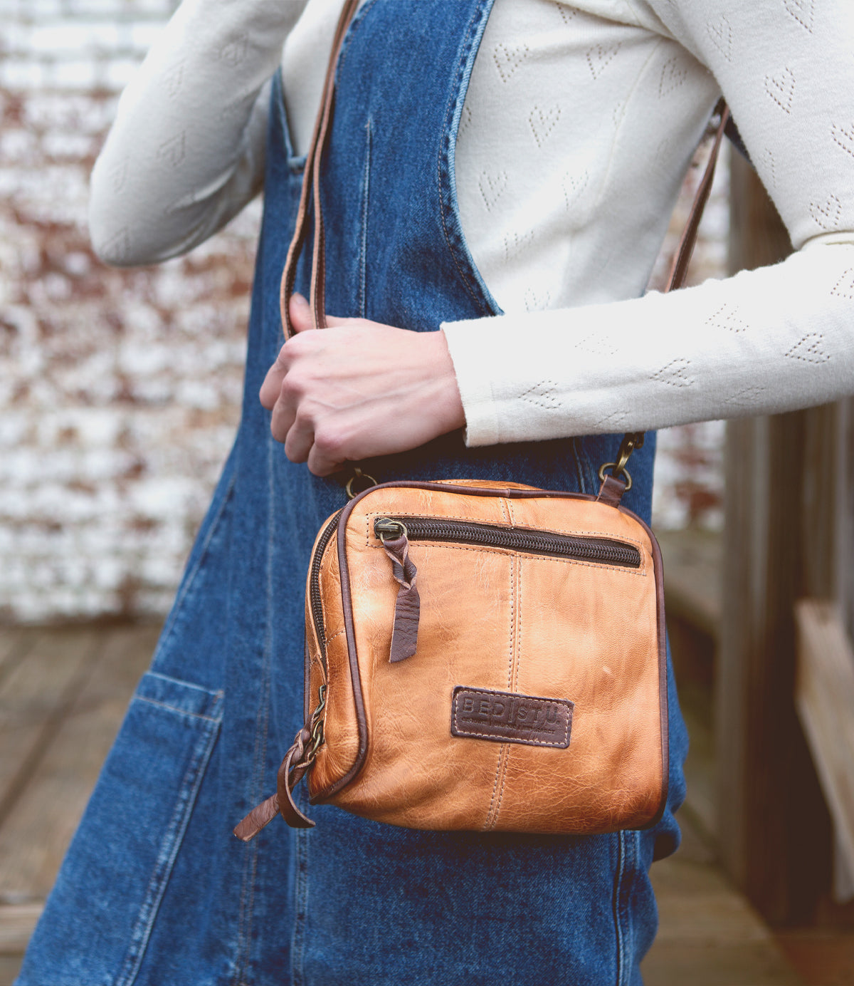 A person in a white long-sleeve top and blue denim pinafore holds the Bed|Stü Capture compact handbag in tan, featuring a front zipper pocket and embossed patch.