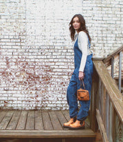 Wearing denim overalls and boots, a woman stands on a wooden deck, holding the Bed|Stü Capture handcrafted purse, with a weathered white brick wall in the background.