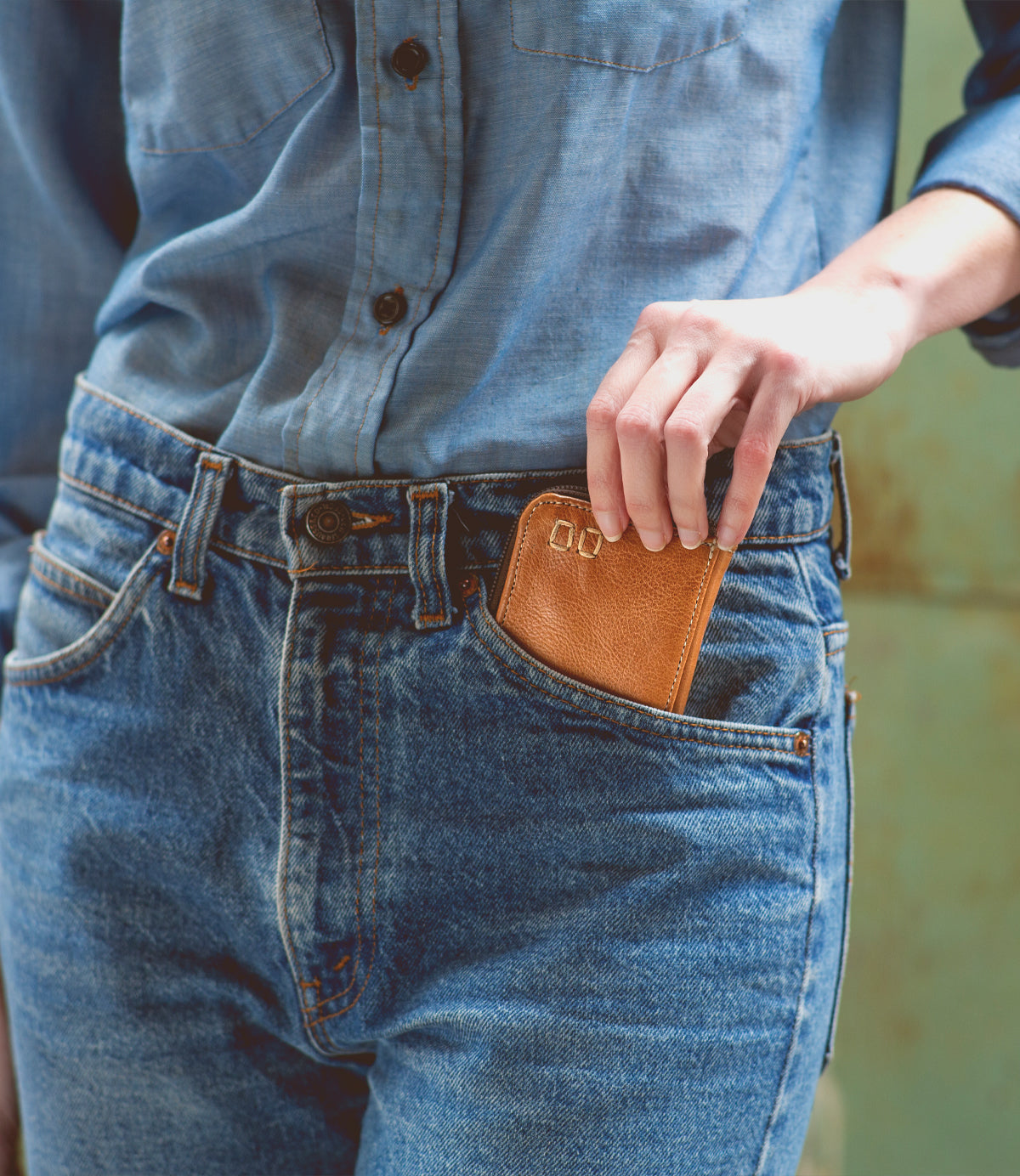 Wearing a blue denim shirt and jeans, the person places the Bed|Stü Carrie compact leather wallet—with card slots and a zip compartment—into their front jeans pocket.