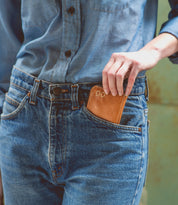 Wearing a blue denim shirt and jeans, the person places the Bed|Stü Carrie compact leather wallet—with card slots and a zip compartment—into their front jeans pocket.