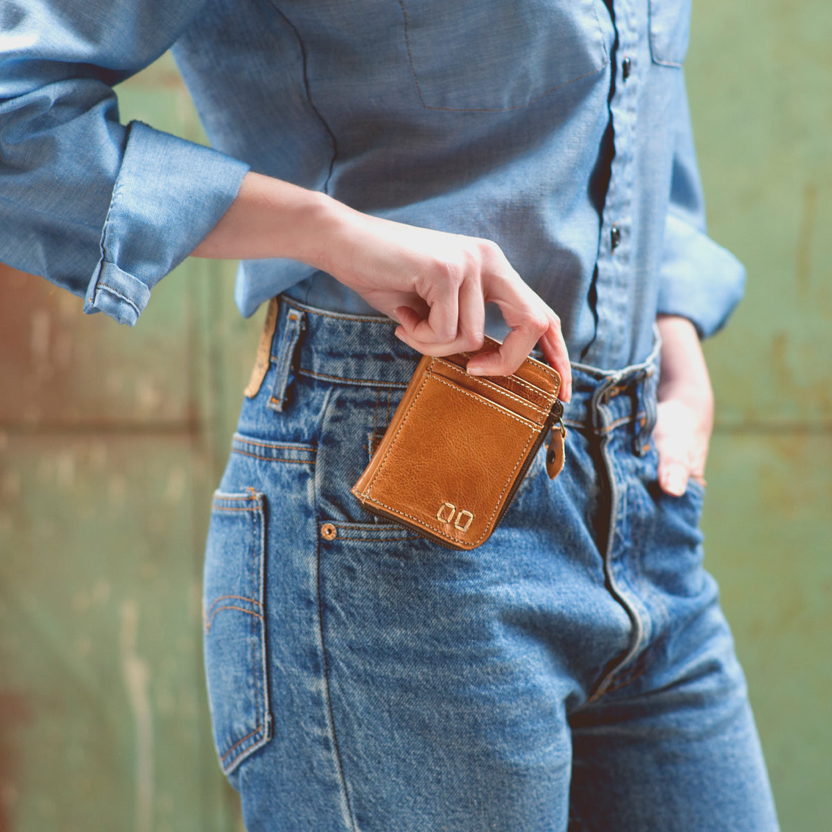 A person in a denim shirt and jeans holds the Bed|Stü Carrie compact leather wallet with one hand in their pocket, the background softly blurred. The Carrie wallet features convenient card slots for easy organization.