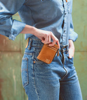 A person in a denim shirt and jeans holds the Bed|Stü Carrie compact leather wallet with one hand in their pocket, the background softly blurred. The Carrie wallet features convenient card slots for easy organization.