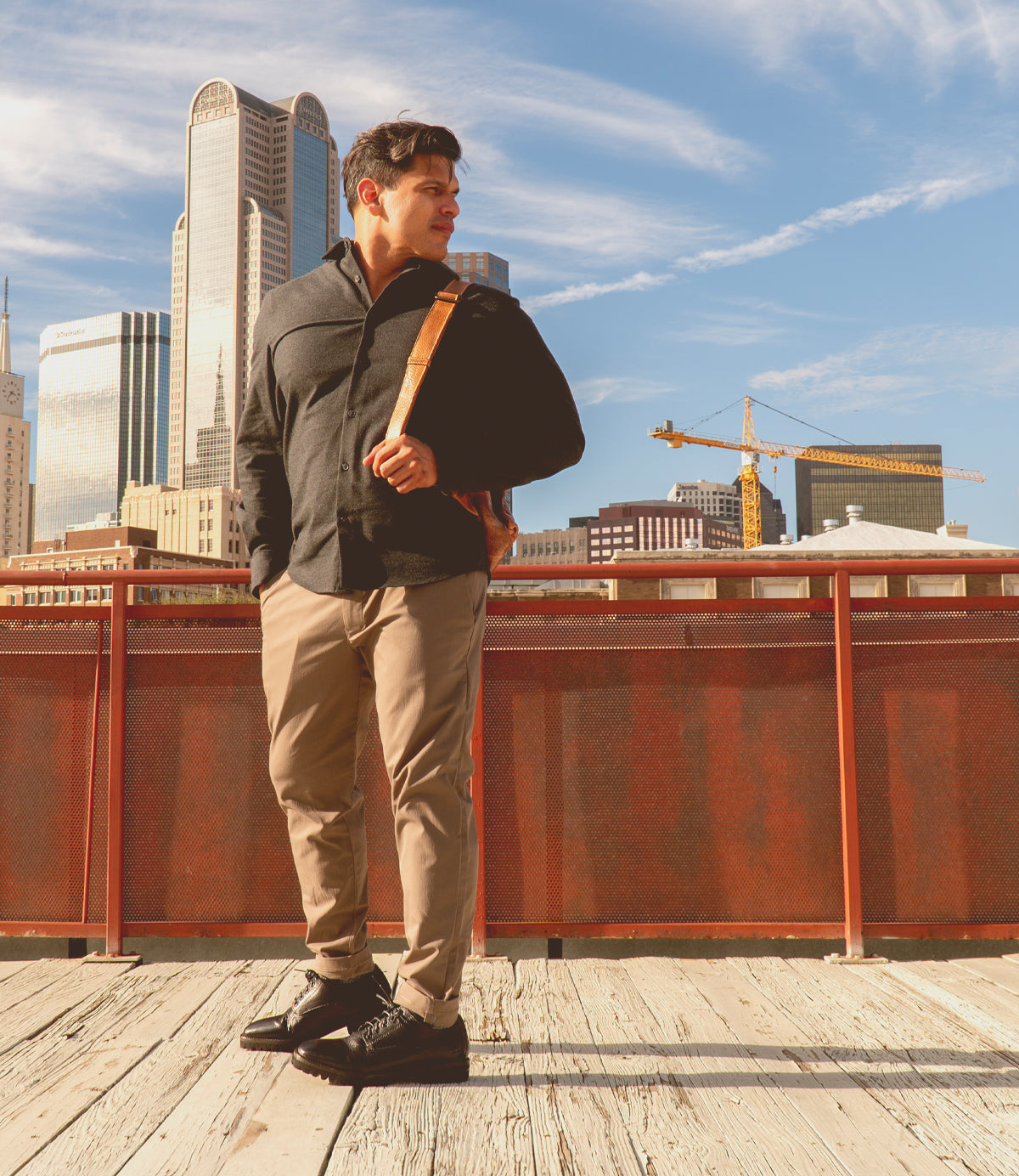 A man stands on a wooden platform in front of a red railing, wearing the Carter oxford dress shoes by Bed|Stü, featuring Goodyear welt construction and a rugged outsole. City buildings and construction cranes are visible in the background under a blue sky.
