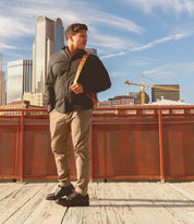 A man stands on a wooden platform in front of a red railing, wearing the Carter oxford dress shoes by Bed|Stü, featuring Goodyear welt construction and a rugged outsole. City buildings and construction cranes are visible in the background under a blue sky.