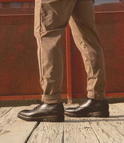 A person wearing tan pants and black Bed|Stü Carter leather oxford dress shoes with Goodyear welt construction stands on a wooden surface with a red metal fence in the background.