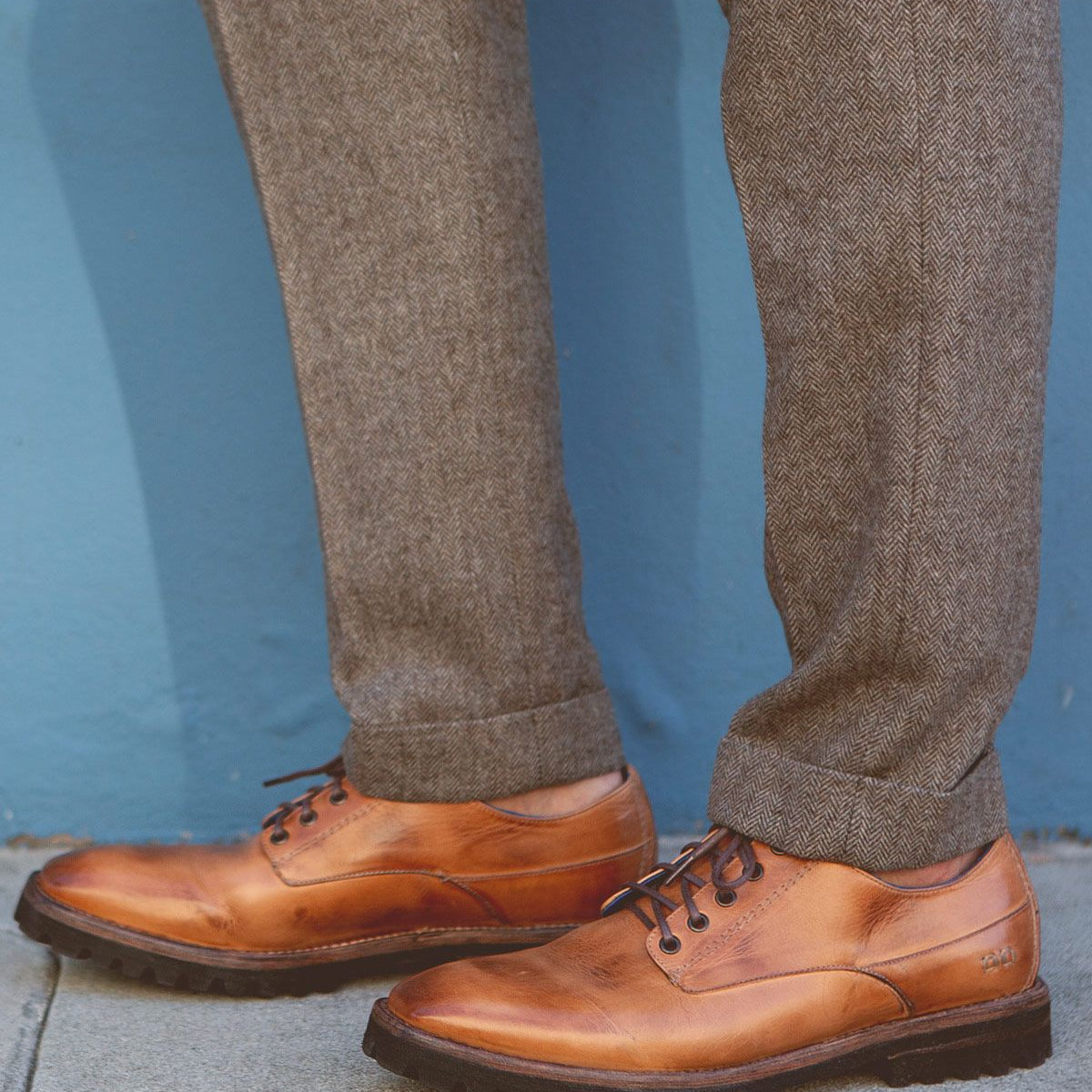 A person wearing the Bed|Stü Carter brown leather oxford dress shoes with a rugged outsole and grey herringbone trousers, standing on a sidewalk against a blue wall.