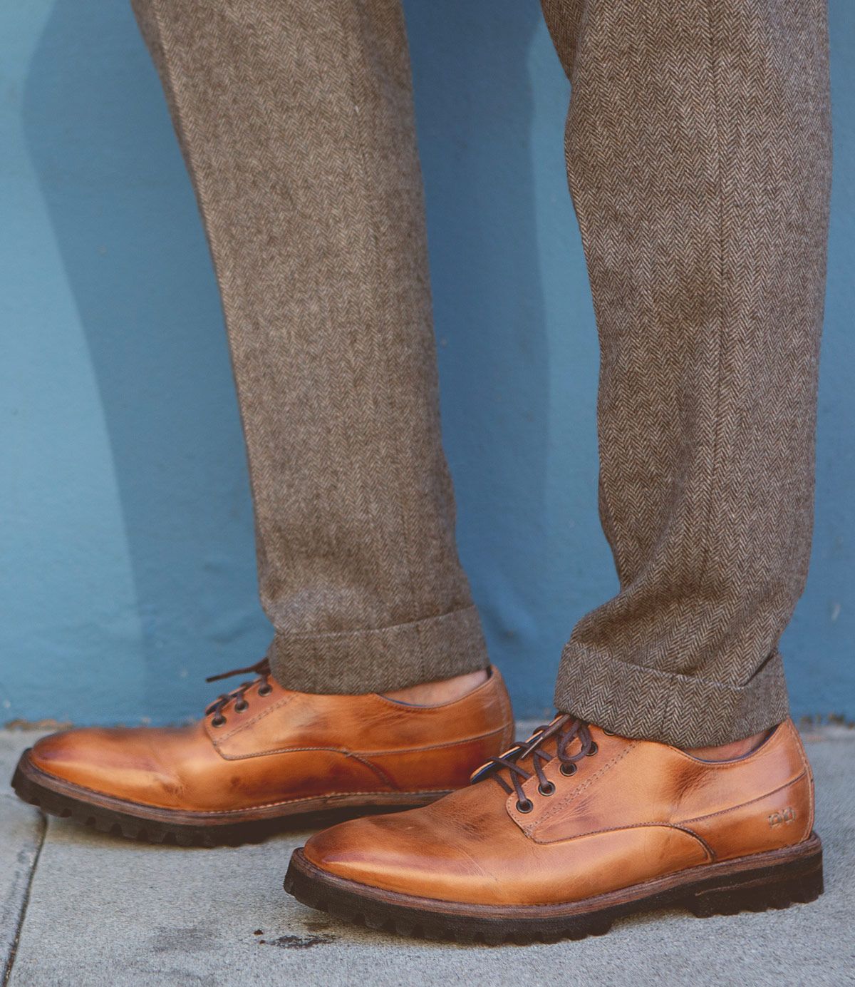 A person wearing the Bed|Stü Carter brown leather oxford dress shoes with a rugged outsole and grey herringbone trousers, standing on a sidewalk against a blue wall.