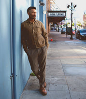 A man in a brown jacket and gray pants leans against a blue wall on a city sidewalk, effortlessly showcasing classic style with the Carter oxford dress shoes by Bed|Stü, featuring Goodyear welt construction. A "Now Playing" marquee and street lamps illuminate the background.