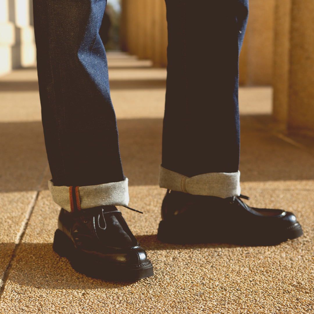 Wearing cuffed dark blue jeans and black vegetable-tanned leather Casotto men’s moc-toe boots by Bed|Stü, the person stands on a sunlit textured sidewalk near tan columns, showcasing timeless Italian craftsmanship.