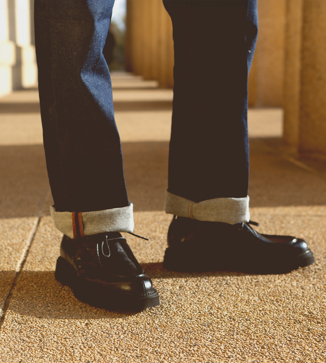 Wearing cuffed dark blue jeans and black vegetable-tanned leather Casotto men’s moc-toe boots by Bed|Stü, the person stands on a sunlit textured sidewalk near tan columns, showcasing timeless Italian craftsmanship.