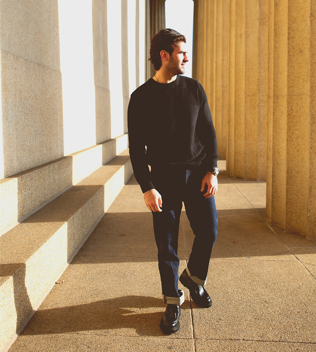 A man wearing a black sweater and jeans walks between large stone columns in bright sunlight, his Bed|Stü Casotto men’s moc-toe boots—crafted with Italian craftsmanship from vegetable-tanned leather—casting distinct shadows on the ground.