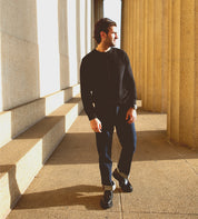 A man wearing a black sweater and jeans walks between large stone columns in bright sunlight, his Bed|Stü Casotto men’s moc-toe boots—crafted with Italian craftsmanship from vegetable-tanned leather—casting distinct shadows on the ground.