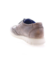 The Bed|Stü Cayuga II is a brown leather brogue shoe featuring classic brogue detailing and a white sole, shown from the back left against a white background.