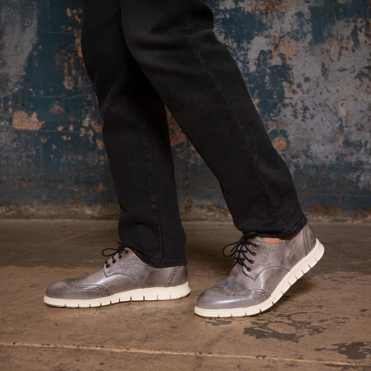 A person wearing black pants and Bed|Stü's Cayuga II silver-gray wingtip shoes with brogue detailing stands on a worn concrete floor against a distressed blue wall.