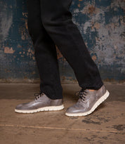 A person wearing black pants and Bed|Stü's Cayuga II silver-gray wingtip shoes with brogue detailing stands on a worn concrete floor against a distressed blue wall.