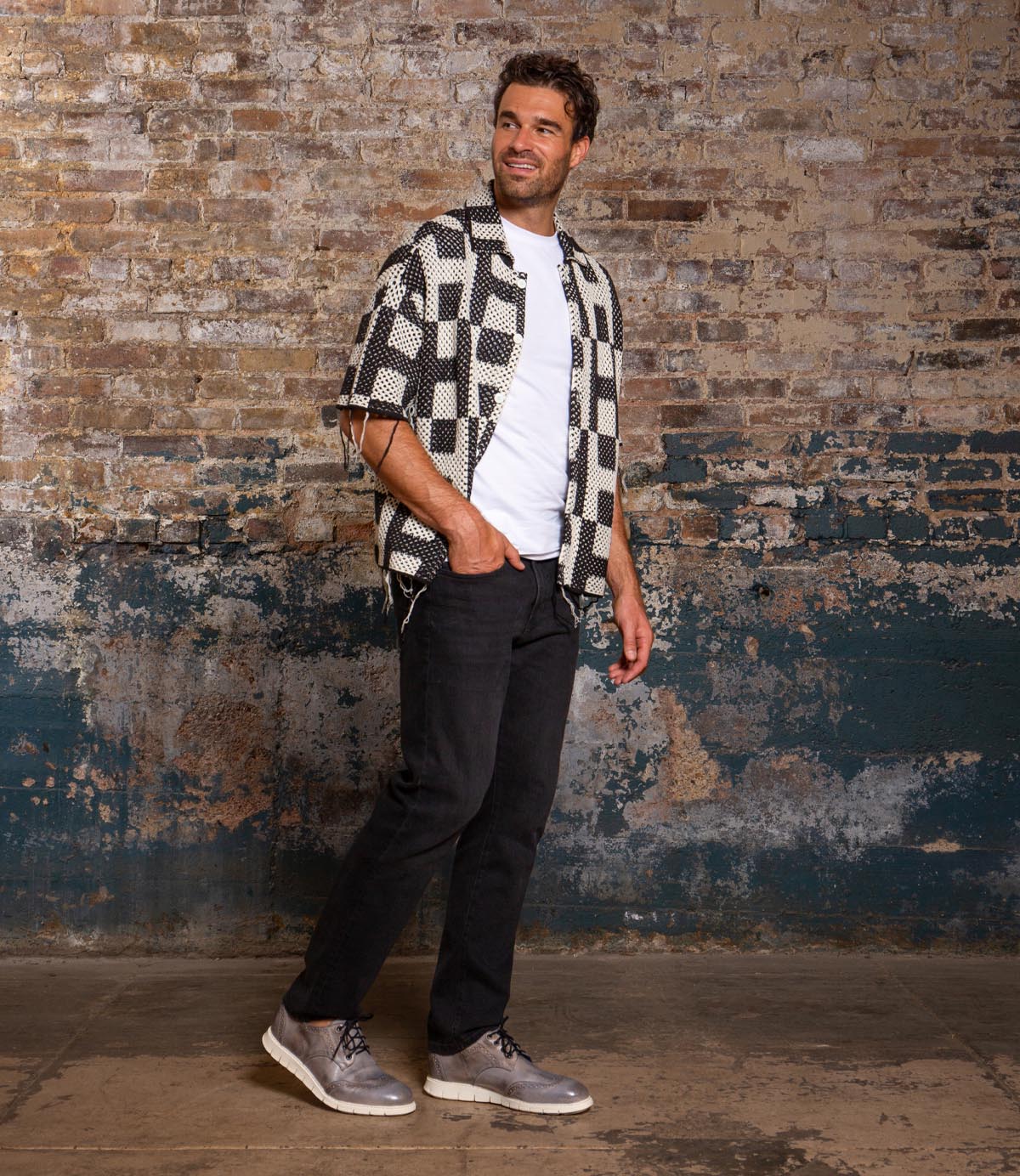 A man stands indoors against a weathered brick wall, wearing a patterned short-sleeve shirt over a white t-shirt, black pants, and gray Bed|Stü Cayuga II derby shoes with subtle brogue detailing.