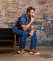 A man in a blue shirt and jeans sits on the edge of a black leather sofa, smiling with his hand on his chin against a weathered brick wall. His Bed|Stü Cayuga II derby shoes, featuring classic brogue detailing, add a polished touch to the relaxed scene.