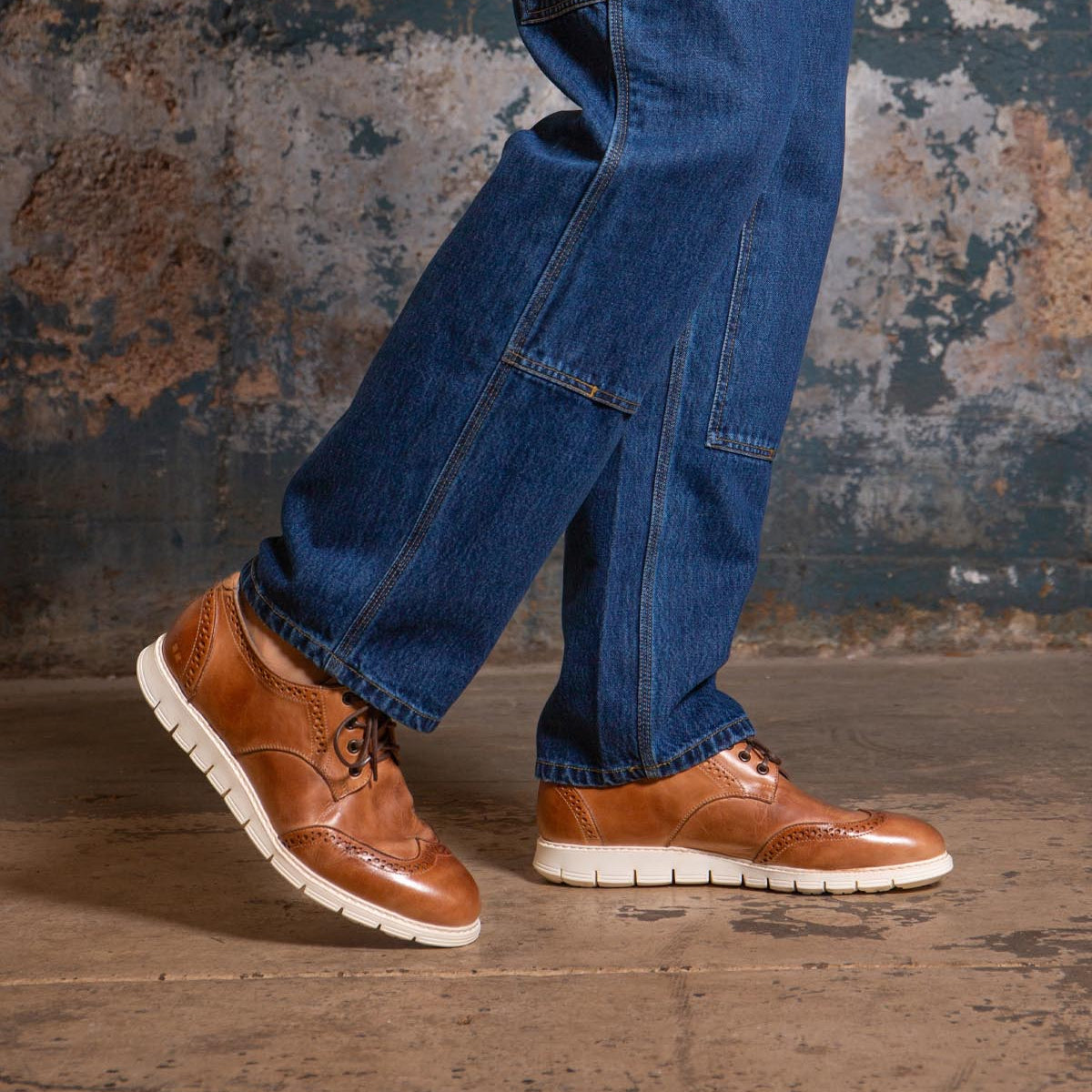 A person wearing blue jeans and the Bed|Stü Cayuga II brown leather derby shoes with brogue detailing and white soles stands on a concrete floor in front of a worn, textured wall.