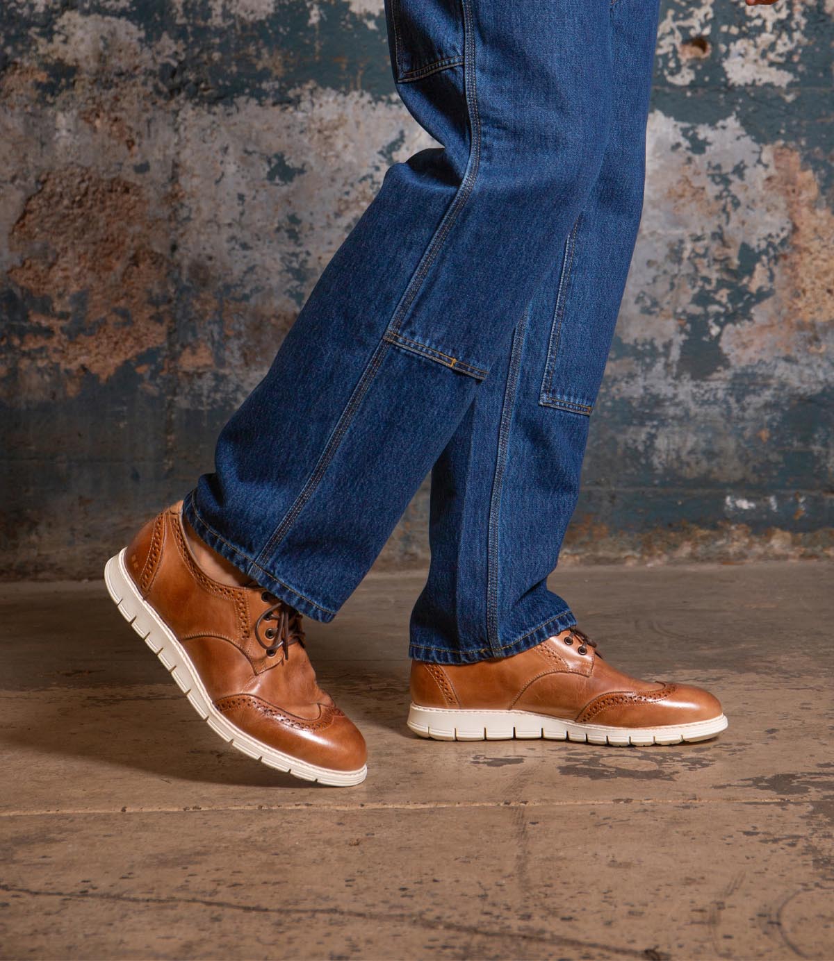 A person wearing blue jeans and the Bed|Stü Cayuga II brown leather derby shoes with brogue detailing and white soles stands on a concrete floor in front of a worn, textured wall.