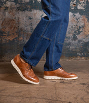 A person wearing blue jeans and the Bed|Stü Cayuga II brown leather derby shoes with brogue detailing and white soles stands on a concrete floor in front of a worn, textured wall.