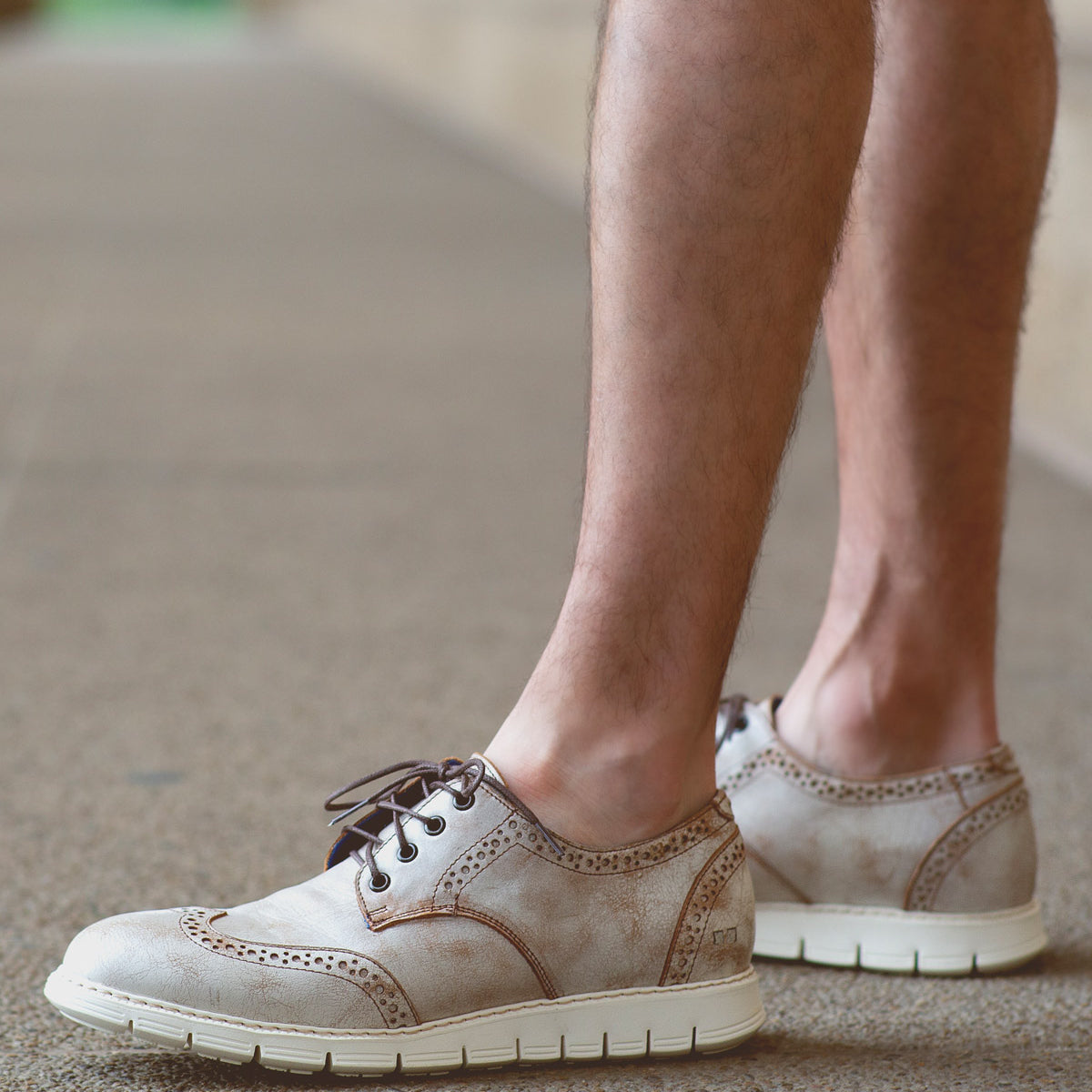 A person wearing the Bed|Stü Cayuga II beige lace-up derby shoes with brogue detailing and white soles stands on a textured outdoor surface.