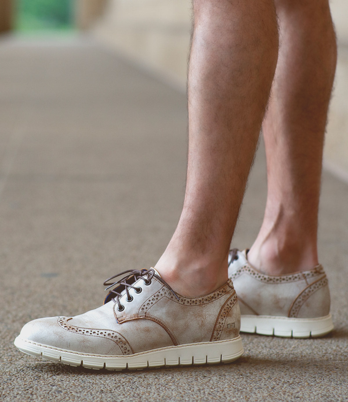 A person wearing the Bed|Stü Cayuga II beige lace-up derby shoes with brogue detailing and white soles stands on a textured outdoor surface.