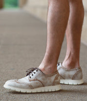 A person wearing the Bed|Stü Cayuga II beige lace-up derby shoes with brogue detailing and white soles stands on a textured outdoor surface.