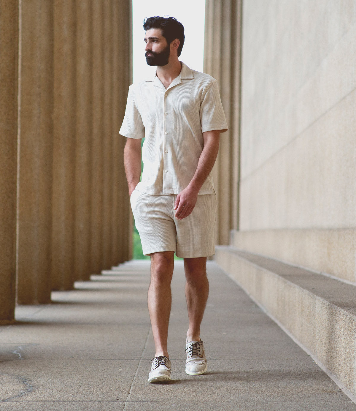 A man with a beard walks along a columned corridor wearing a light short-sleeve button-up shirt, matching shorts, and Bed|Stü’s Cayuga II casual sneakers featuring subtle brogue detailing.
