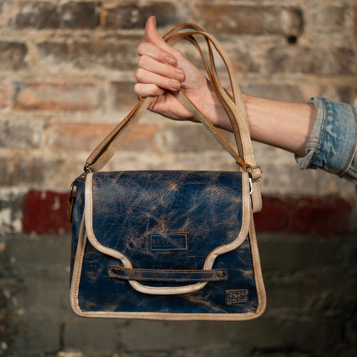 A hand holds the Bed|Stü Ceremonious, a distressed blue leather handbag with beige trim and a long strap, showcasing its preppy flap-top design and secure magnetic closure, against a brick wall.