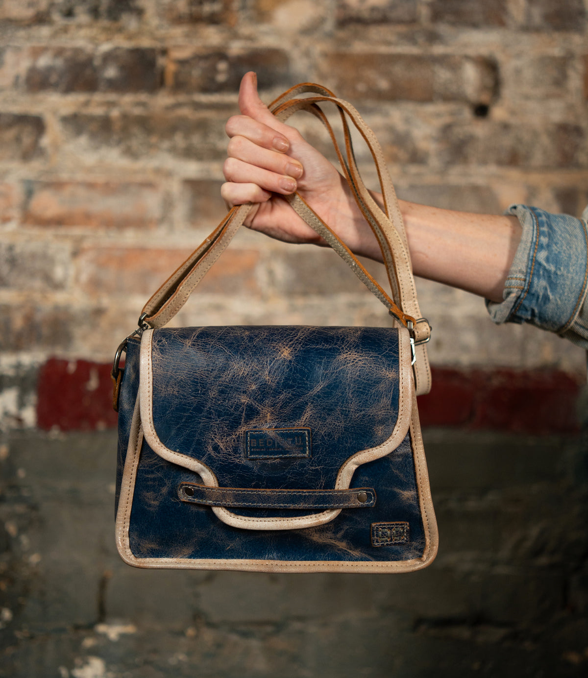 A hand holds the Bed|Stü Ceremonious, a distressed blue leather handbag with beige trim and a long strap, showcasing its preppy flap-top design and secure magnetic closure, against a brick wall.