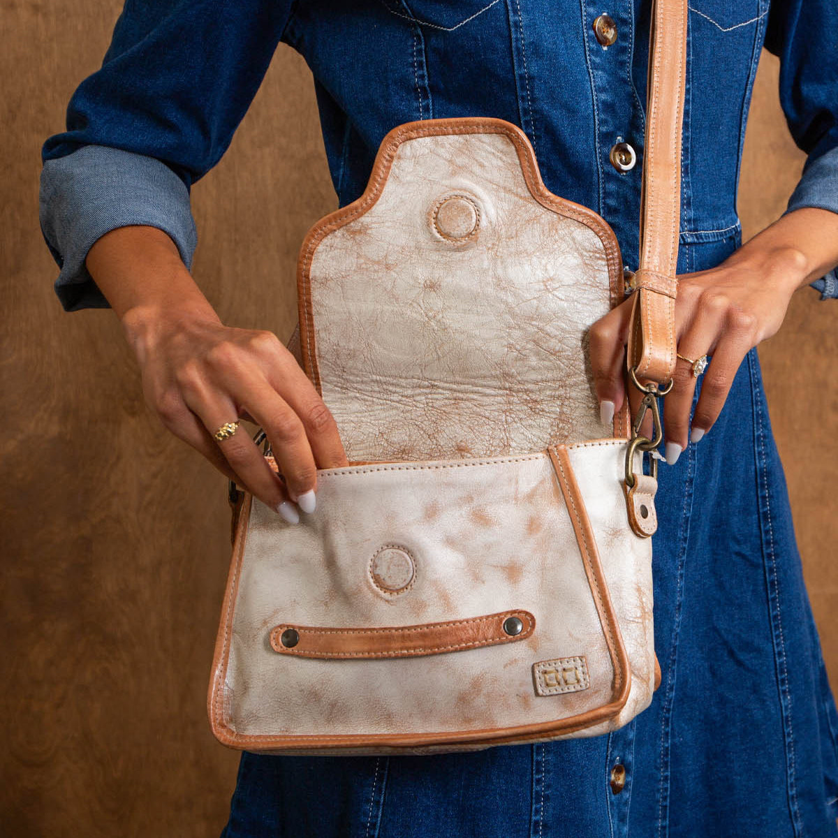 A person wearing a blue denim dress holds an open Bed|Stü Ceremonious beige and tan leather preppy flap-top bag with a magnetic closure, showing its organized interior against a brown background.