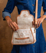 A person wearing a blue denim dress holds an open Bed|Stü Ceremonious beige and tan leather preppy flap-top bag with a magnetic closure, showing its organized interior against a brown background.