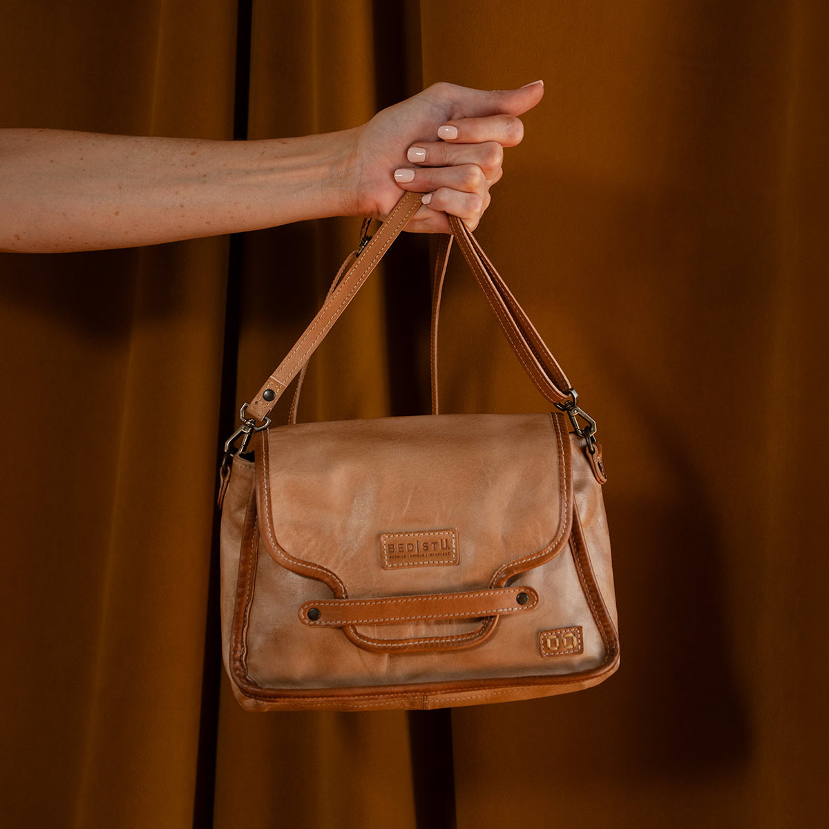 A hand holds the Bed|Stü Ceremonious flap-top bag, expertly crafted from tan leather with stitched accents, a magnetic closure, an organized interior, and a subtle logo patch—all set against a brown backdrop.