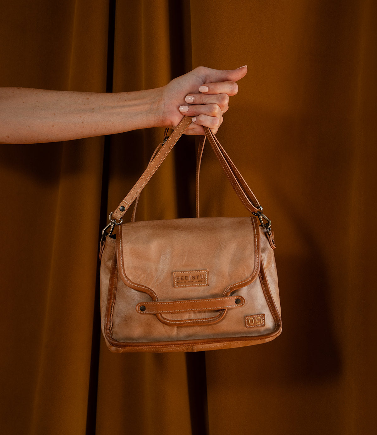A hand holds the Bed|Stü Ceremonious flap-top bag, expertly crafted from tan leather with stitched accents, a magnetic closure, an organized interior, and a subtle logo patch—all set against a brown backdrop.