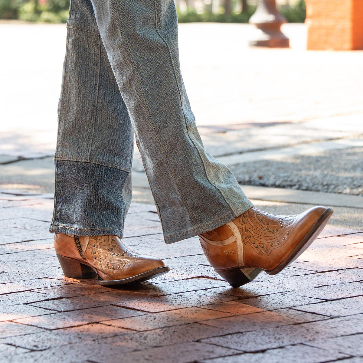 A person wearing the Bed|Stü Chamise mid-height western boots in light brown with blue jeans, walking on a brick sidewalk outdoors.