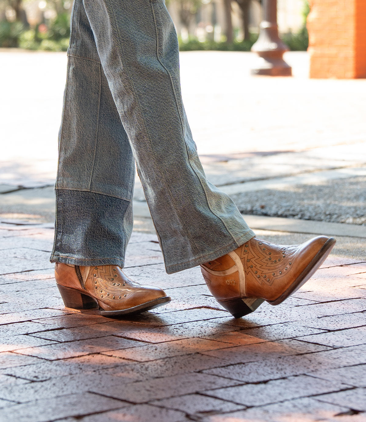 A person wearing the Bed|Stü Chamise mid-height western boots in light brown with blue jeans, walking on a brick sidewalk outdoors.