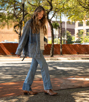 A woman walks outdoors on a sunny day wearing Bed|Stü's Chamise brown mid-height boots, paired with a denim jacket, flared jeans, and a beige top with fringe. Trees and buildings are visible in the background.