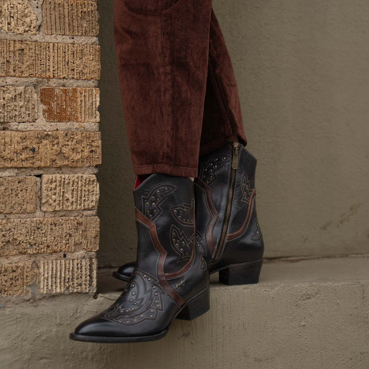 A person wearing Bed|Stü’s Chamise dark, embroidered western boots and brown corduroy pants stands on a concrete ledge next to a brick wall.