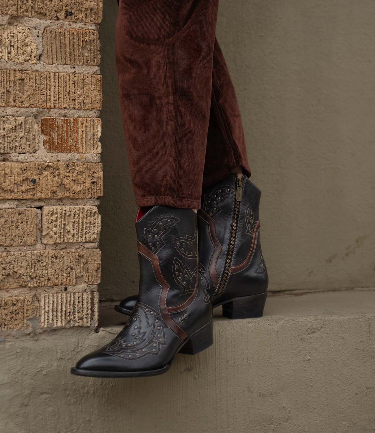 A person wearing Bed|Stü’s Chamise dark, embroidered western boots and brown corduroy pants stands on a concrete ledge next to a brick wall.